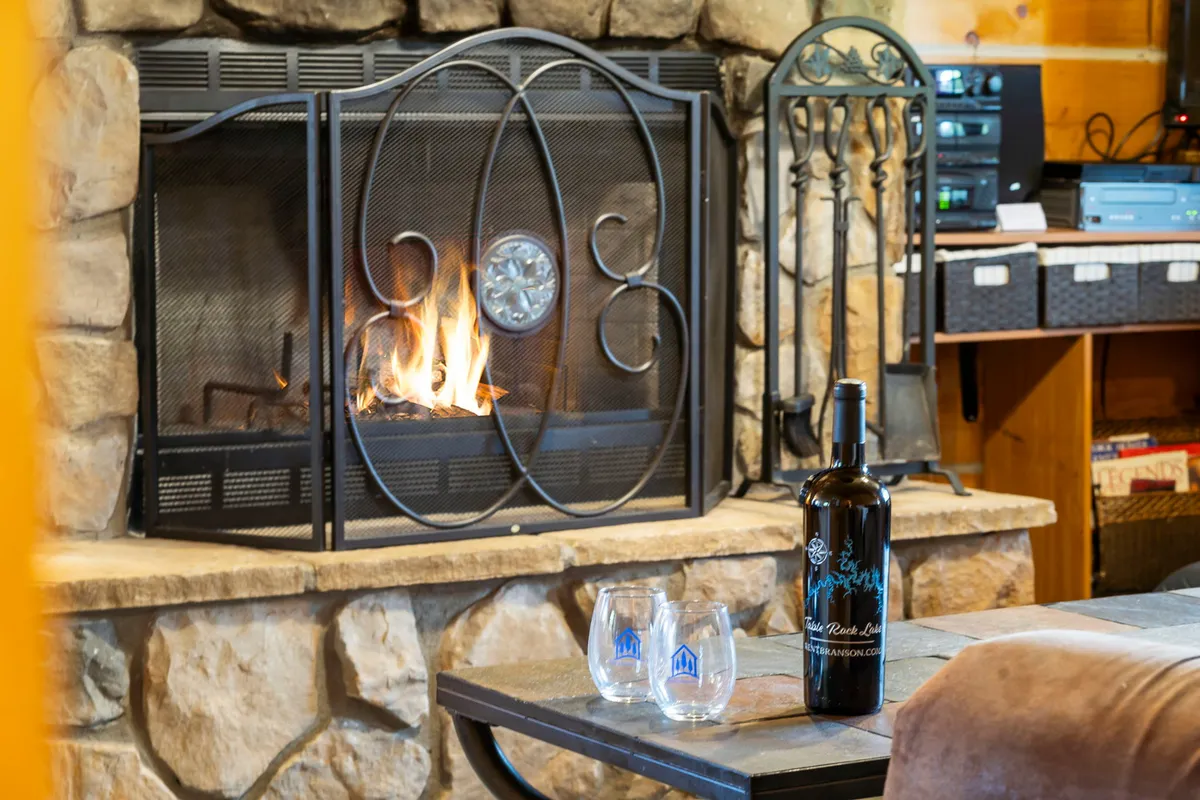 A close-up of a cozy fireplace scene, featuring a bottle of wine and glasses on the hearth, surrounded by a warm glow. The setting suggests intimate moments, relaxation, and special celebrations—ideal for couples or friends seeking a getaway in a safe, clean cabin environment.