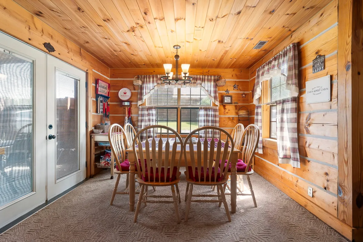 A spacious wooden dining table sits under a glowing chandelier, surrounded by large windows that let in natural light. Warm wooden walls frame the scene, evoking a sense of togetherness and comfort perfect for group gatherings or shared meals.