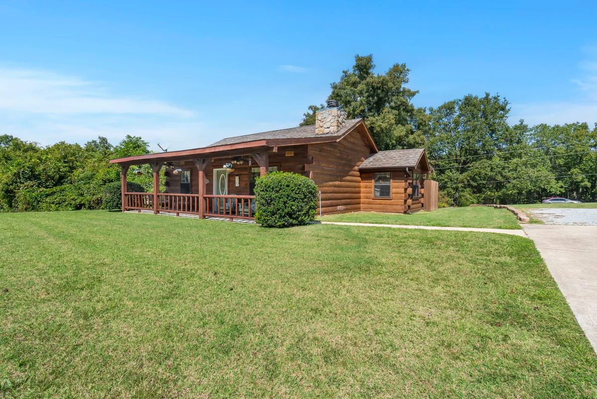 A classic log cabin features a welcoming front porch with chairs perfect for relaxing, set amidst a landscaped yard surrounded by lush greenery. The inviting scene suggests a space ideal for family gatherings and moments of relaxation after a day of Ozark adventures.