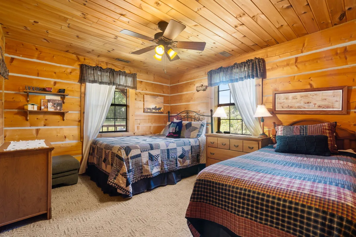 A cozy second bedroom with two beds (a full and a twin), wooden walls, a ceiling fan, and a nightstand with a lamp. The arrangement ensures a welcoming and flexible space for families or friends to relax and connect.