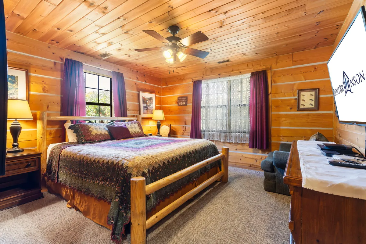A spacious king bedroom featuring a large bed with a decorative quilt, warm wood paneling, a window with curtains letting in natural light, and a TV on the dresser. The room radiates tranquility and comfort, making it ideal for couples or anyone craving a serene retreat.