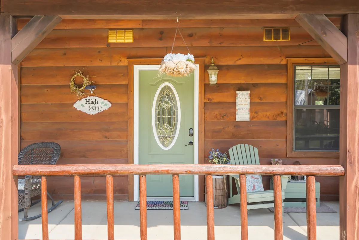 This image shows a green front door adorned with a decorative wreath, a classic rocking chair, and a sign reading 'High Point' on the cozy front porch. The porch is bathed in warm, natural light, creating a welcoming and inviting scene. The image hints at family gatherings, sunset views, and quiet moments spent enjoying the outdoor tranquility.